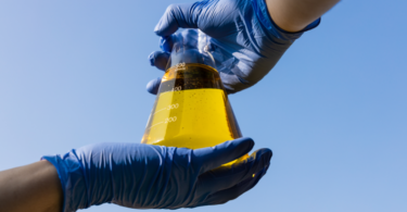 CBIOs: Mão com luvas segurando copo com biocombustível etanol contra o céu azul
