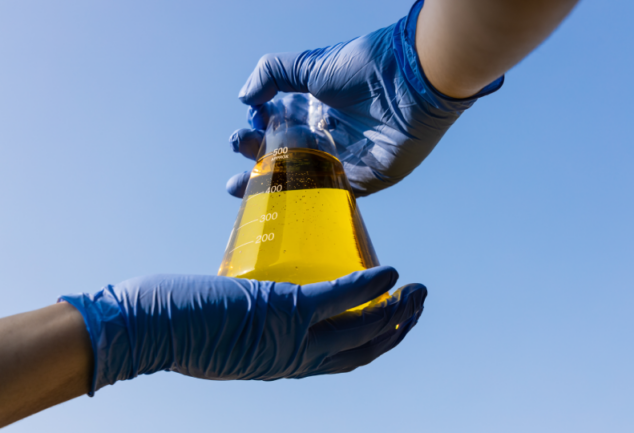 CBIOs: Mão com luvas segurando copo com biocombustível etanol contra o céu azul
